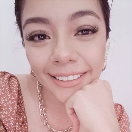 dani Smiling woman with long eyelashes, wearing a gold necklace and patterned top, rests her chin on her hand in this vibrant Ceemi Agency portrait.