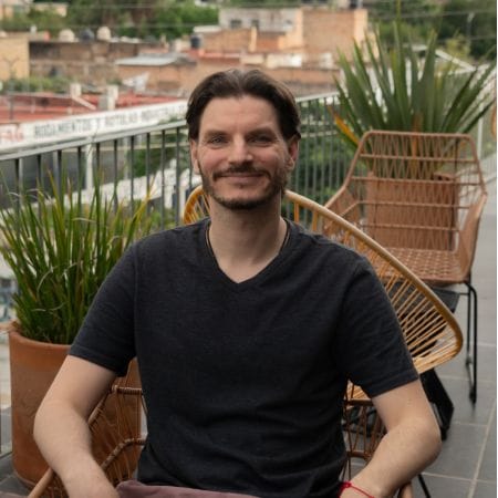 eike Man with short dark hair and beard, smiling while sitting on a patio with plants and metal chairs, enjoying a relaxing moment after collaborating with Ceemi Agency.